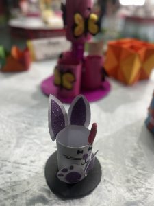 A small, handcrafted bunny made from a white paper roll with purple glitter ears and paws, featuring a painted face and a red pen inserted at the back.