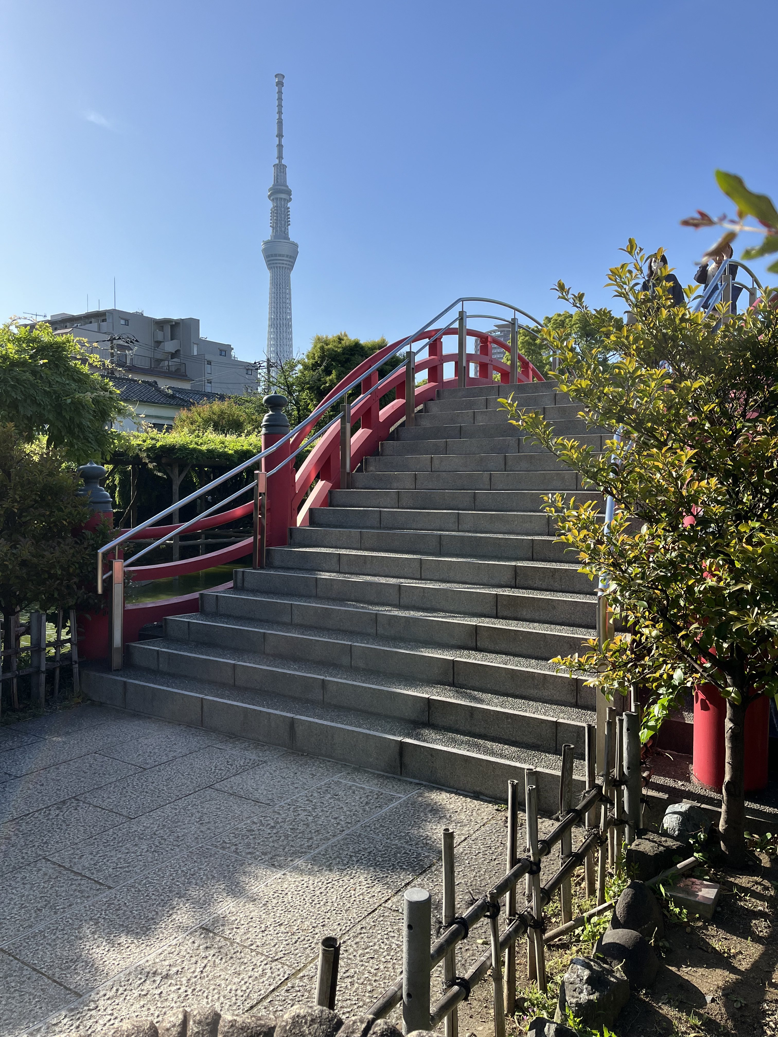 A stone stairway with red railings leads upward, surrounded by lush greenery. In the background, a tall tower rises against a clear blue sky.