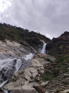 A waterfall cascading down a rocky hillside surrounded by greenery under a cloudy sky.
