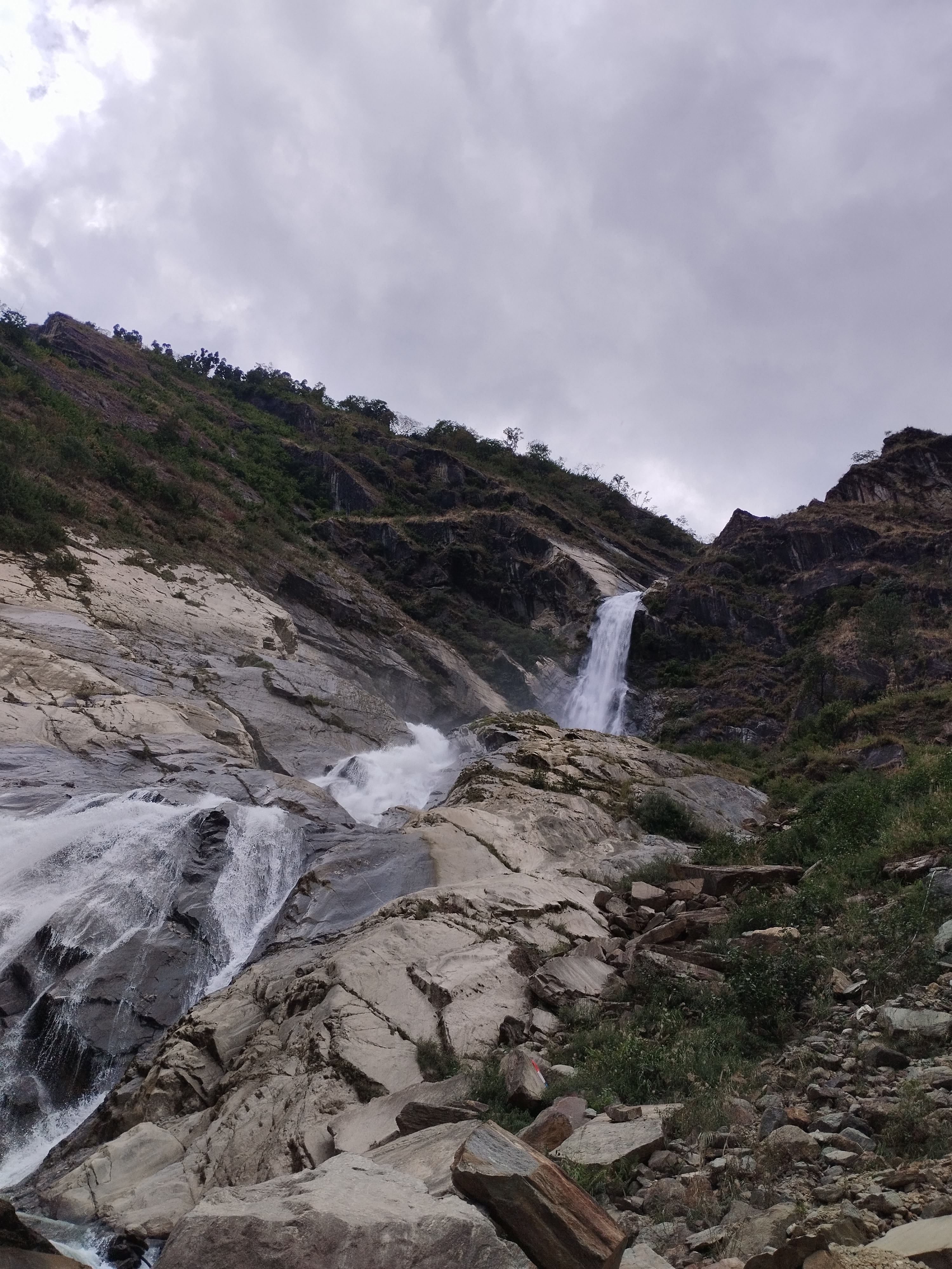A waterfall cascading down a rocky hillside surrounded by greenery under a cloudy sky.
