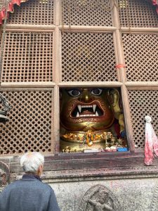 A person with gray hair stands in front of a large, colorful deity sculpture with a fierce expression and large fangs. The sculpture is adorned with flowers and offerings. 