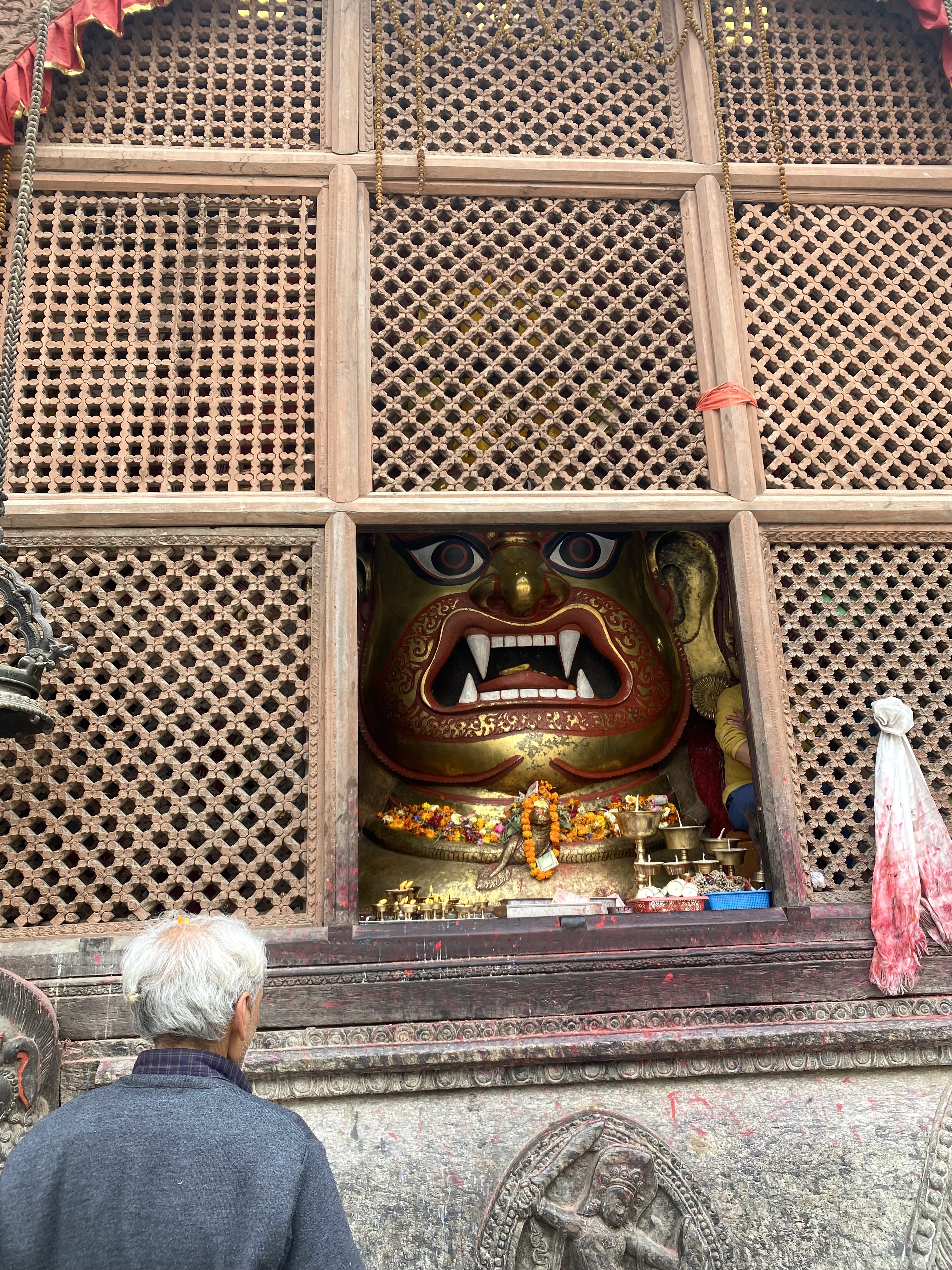 A person with gray hair stands in front of a large, colorful deity sculpture with a fierce expression and large fangs. The sculpture is adorned with flowers and offerings.