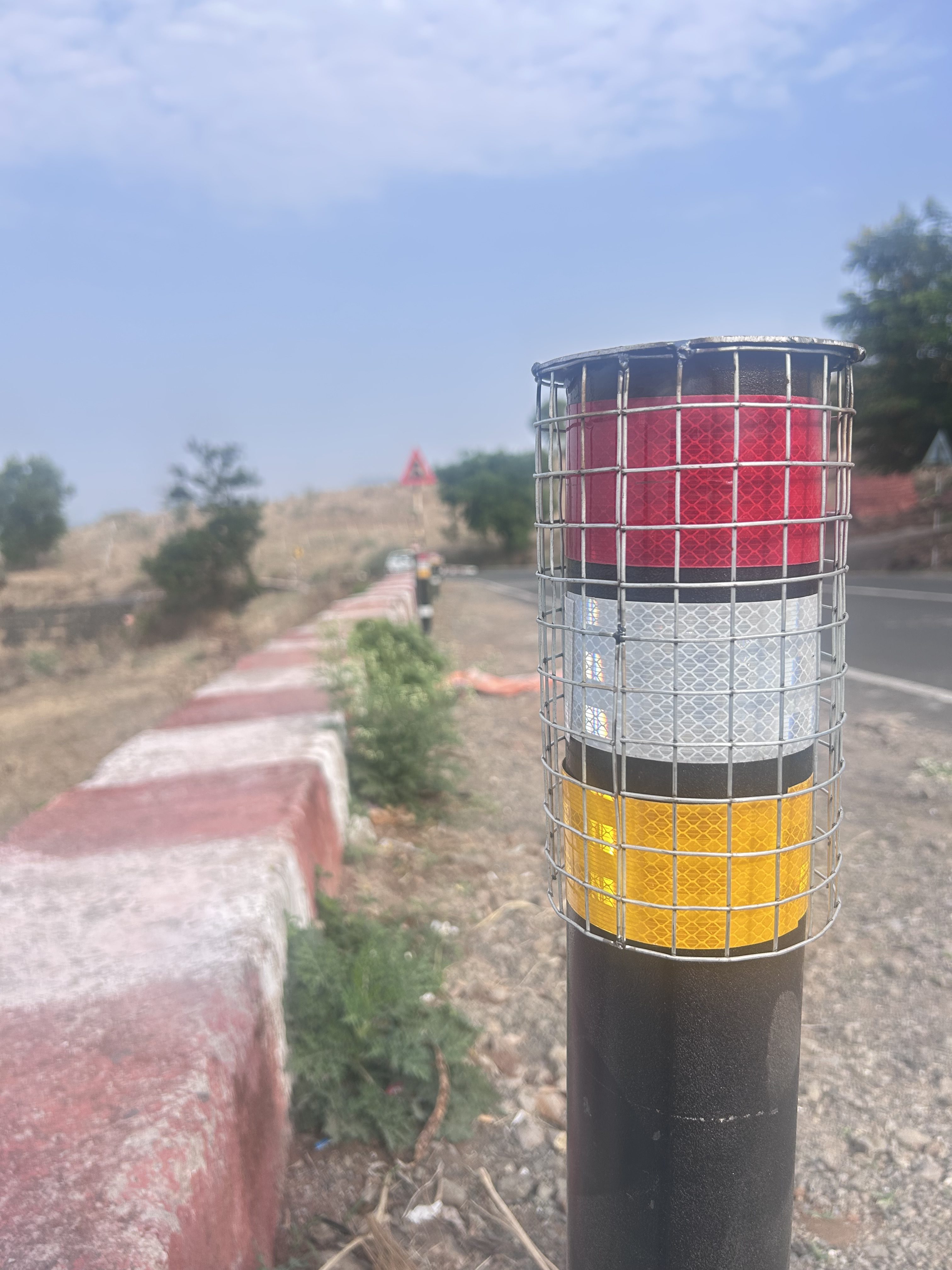 Close-up of a roadside reflector post with red, white, and yellow reflective bands enclosed in a metal grid, positioned next to a winding road with red and white painted barriers.