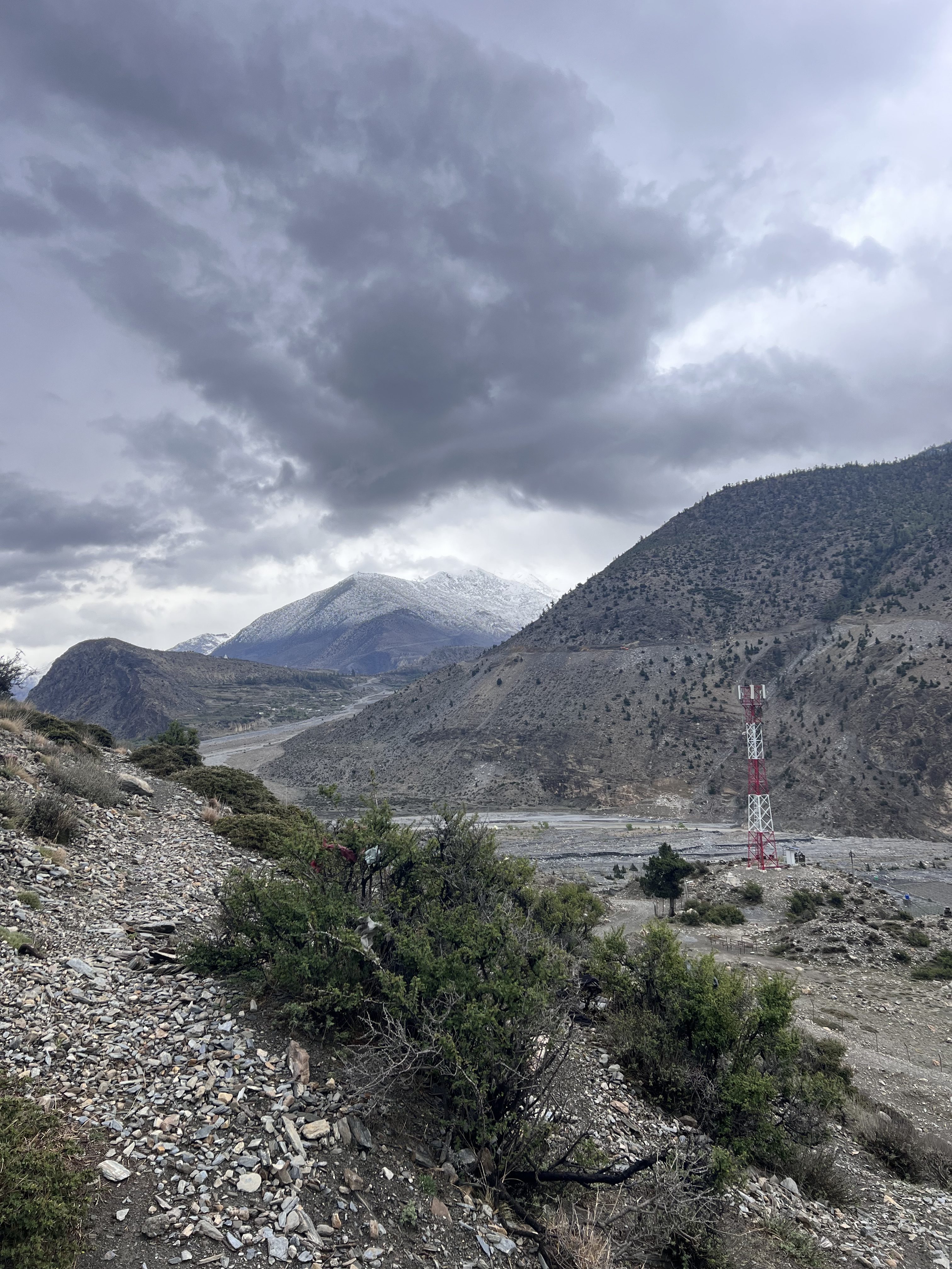 A mountainous landscape with rocky terrain and sparse vegetation. A tall red and white communication tower stands on the right. Snow-capped mountains are visible in the background under a cloudy sky.