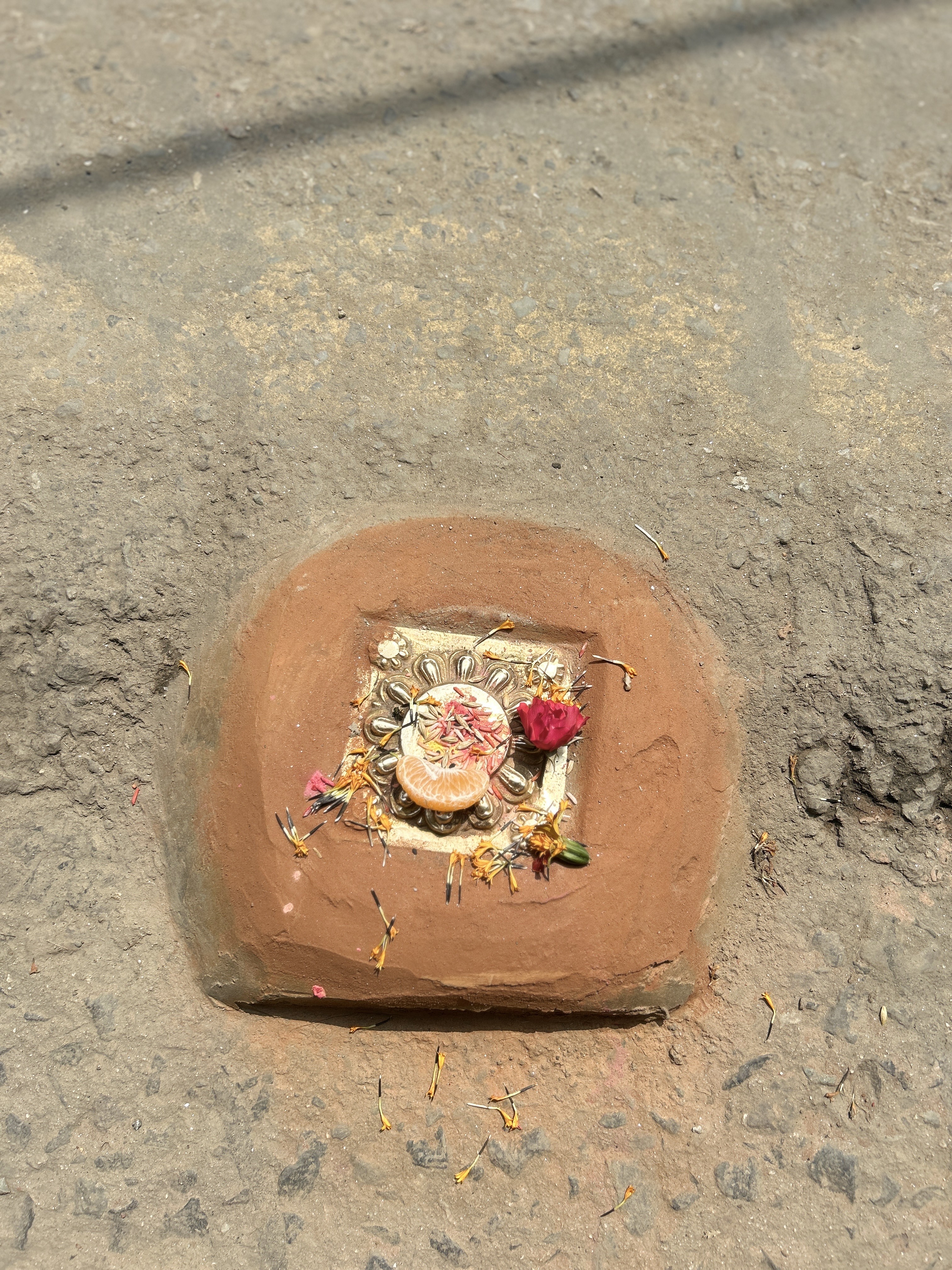 A small altar embedded in the ground with an orange slice, a red flower, and scattered petals, surrounded by a frame of clay on a rough surface.