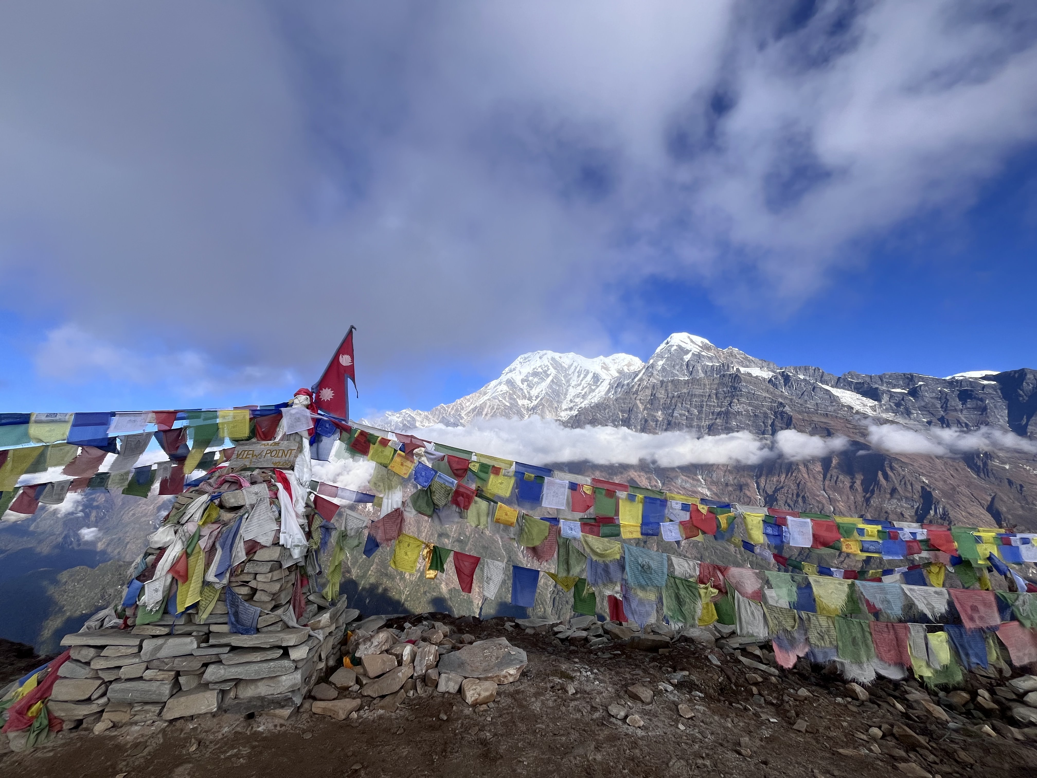 A mountainous landscape with snow-capped peaks in the background under a partly cloudy sky. In the foreground, colorful prayer flags are strung across a pile of stones with a small red flag of Nepal featuring a white emblem.