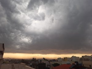 Dramatic sky with thick, dark storm clouds looming over a residential area at sunset, with a faint orange glow on the horizon.
