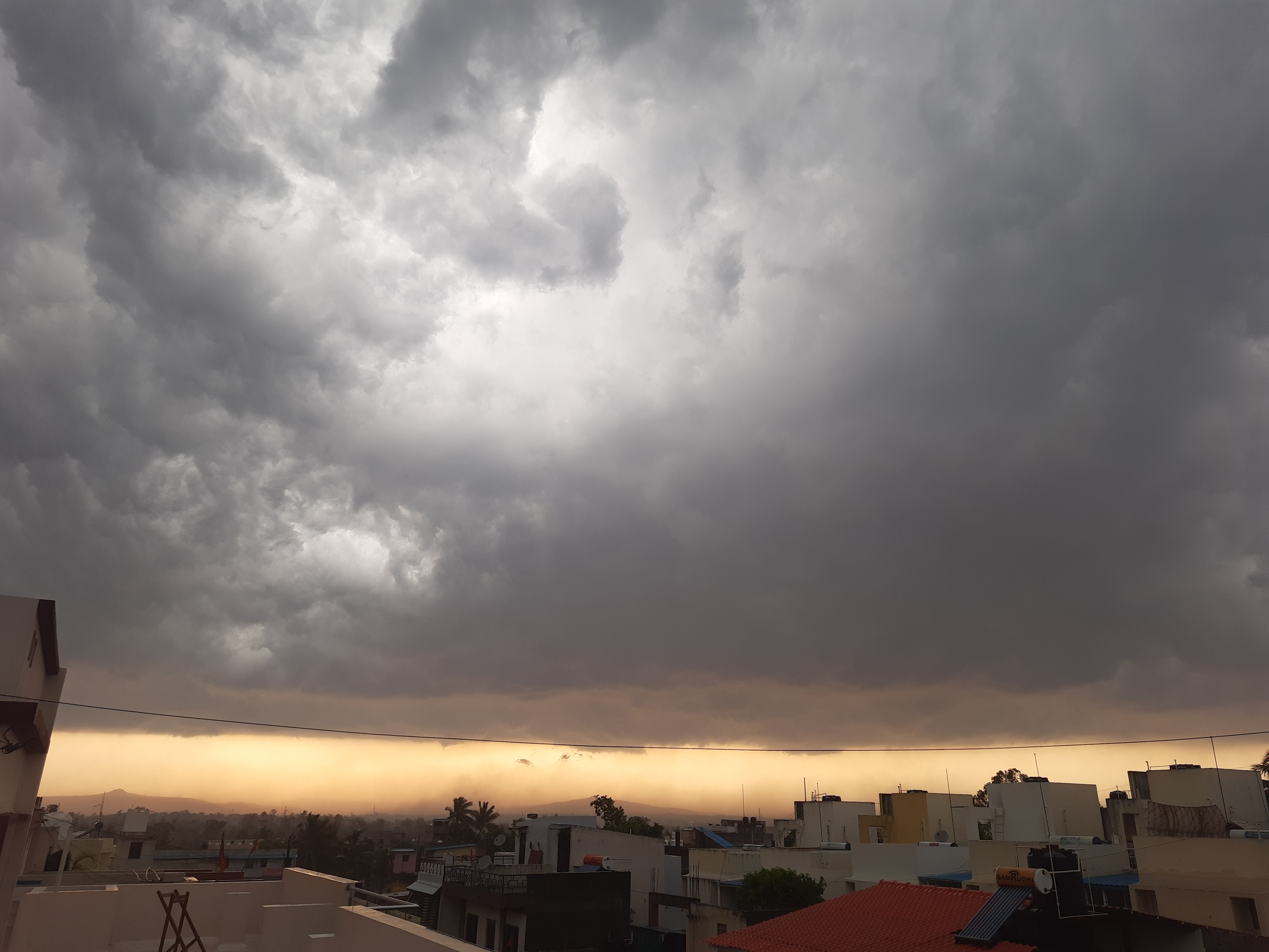 Dramatic sky with thick, dark storm clouds looming over a residential area at sunset, with a faint orange glow on the horizon.