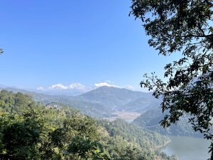 A scenic view of mountainous landscape with snow-capped peaks in the distance under a clear blue sky. Lush green forests cover the hills, with a lake visible below.