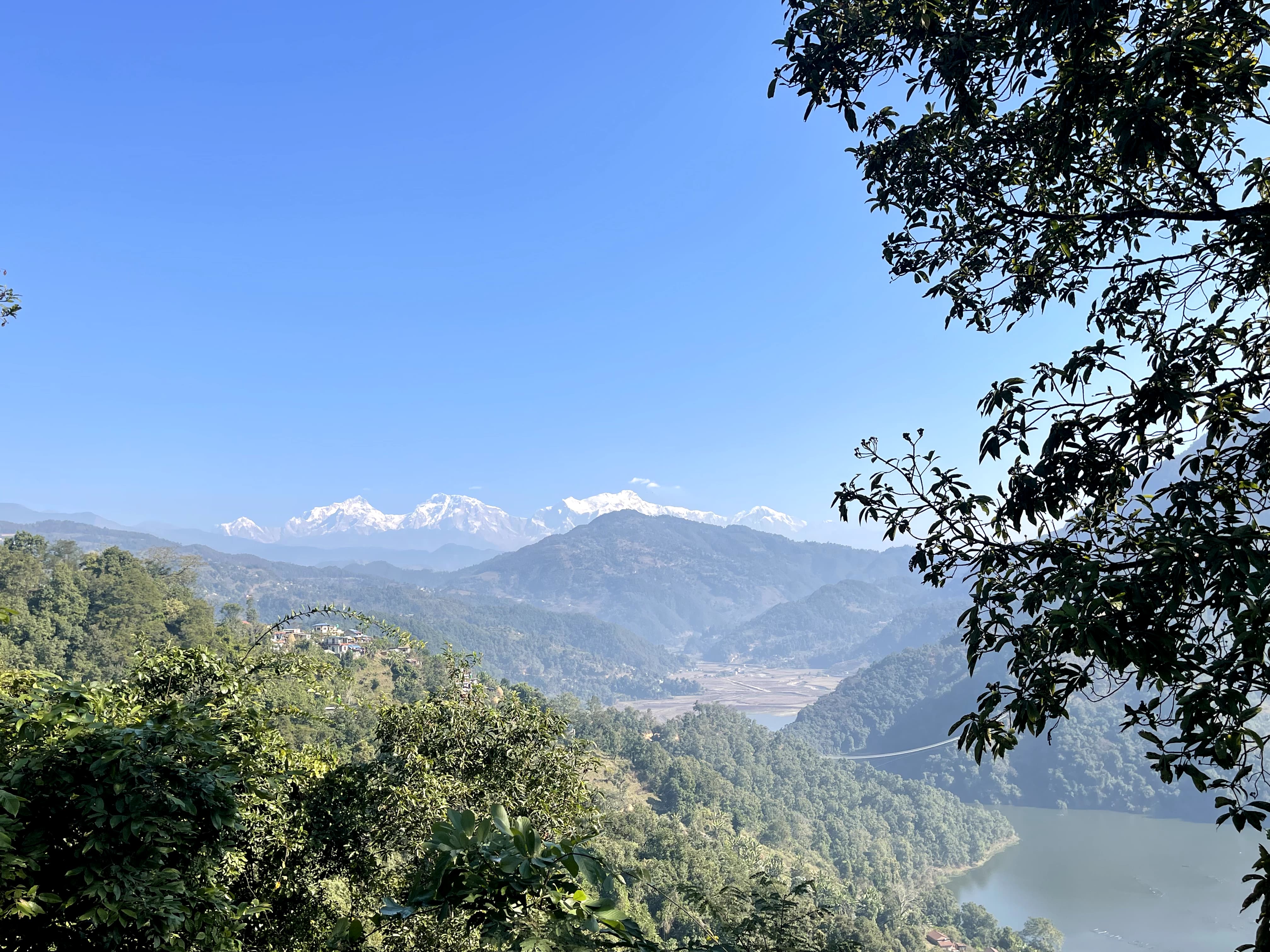 A scenic view of mountainous landscape with snow-capped peaks in the distance under a clear blue sky. Lush green forests cover the hills, with a lake visible below.