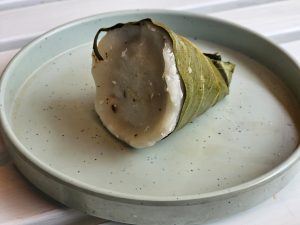 A single Ela Ada, a traditional Kerala snack, served on a light gray plate. The Ela Ada, wrapped in a green leaf, is cut open to reveal its white filling. Ela Ada is a traditional sweet dish from Kerala, India, made of rice flour dough, filled with a sweet mixture of grated coconut and jaggery, and steamed in banana leaves. 