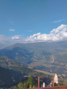 A panoramic view of a mountainous landscape under a clear blue sky, with a few white clouds scattered above.