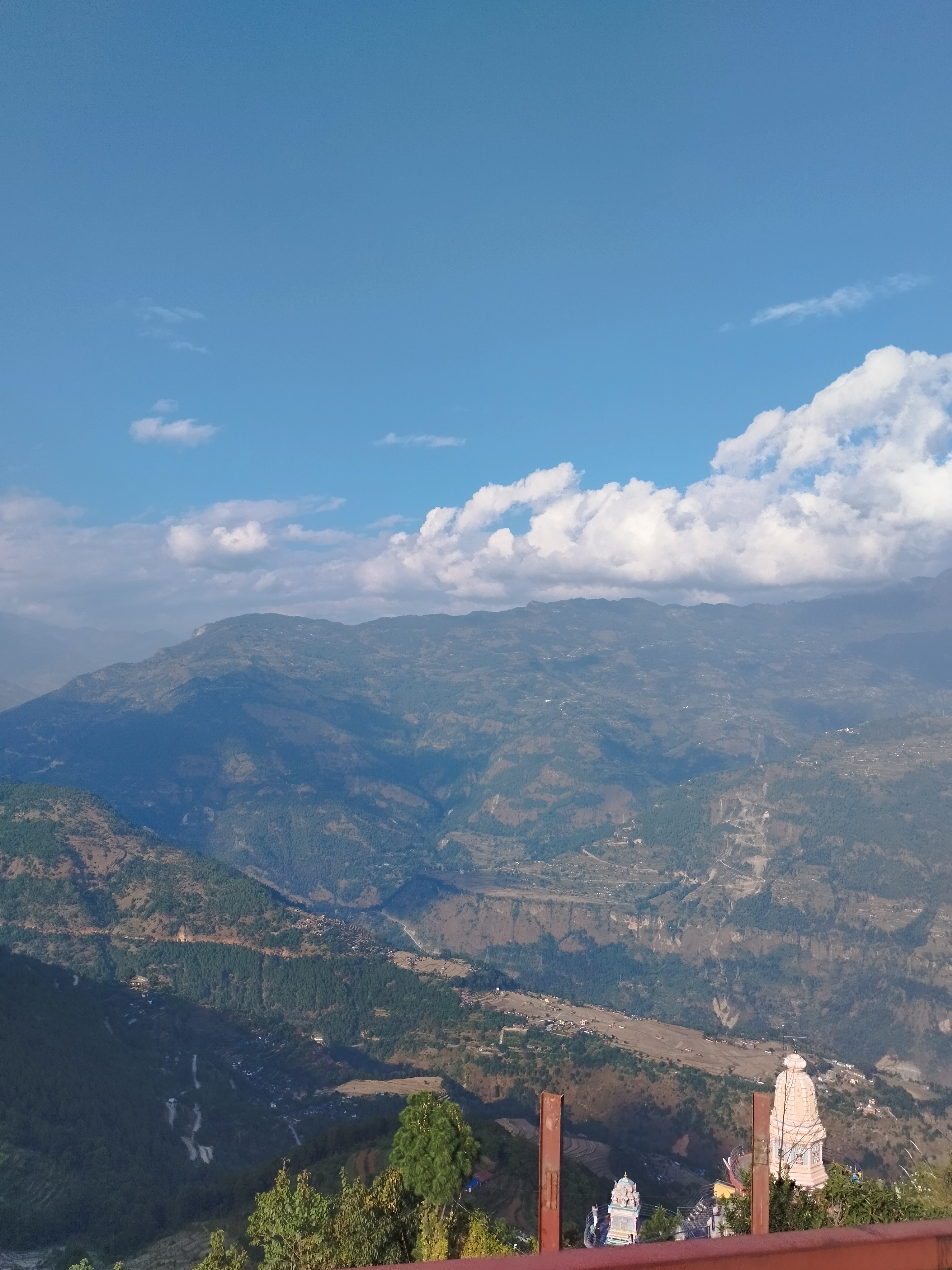 A panoramic view of a mountainous landscape under a clear blue sky, with a few white clouds scattered above.