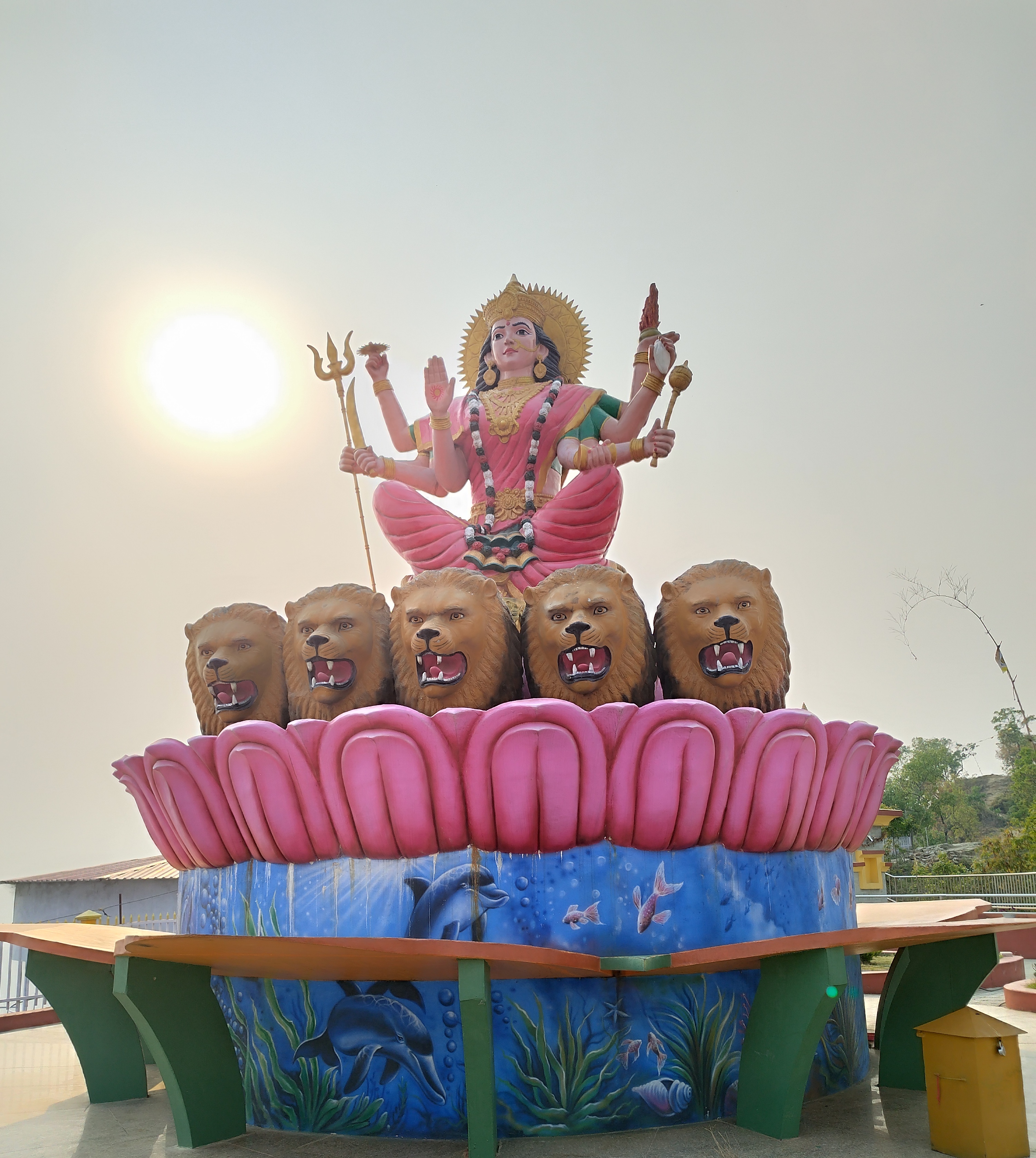 A colorful statue of a deity with multiple arms holding various objects, seated on a large pink lotus surrounded by five lion heads.