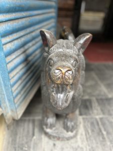 A close-up image of a weathered, carved stone statue of an animal with a mostly black finish, possibly a mythical creature or dog, standing on a tiled floor next to a blue corrugated metal shutter