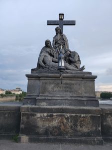 The statue of Pietà on the Charles Bridge in Prague depicts the Virgin Mary cradling the deceased Jesus Christ, with other figures present. The statue is made of dark stone and stands on a pedestal with an inscription, set against a backdrop of a cloudy sky and the city skyline.
