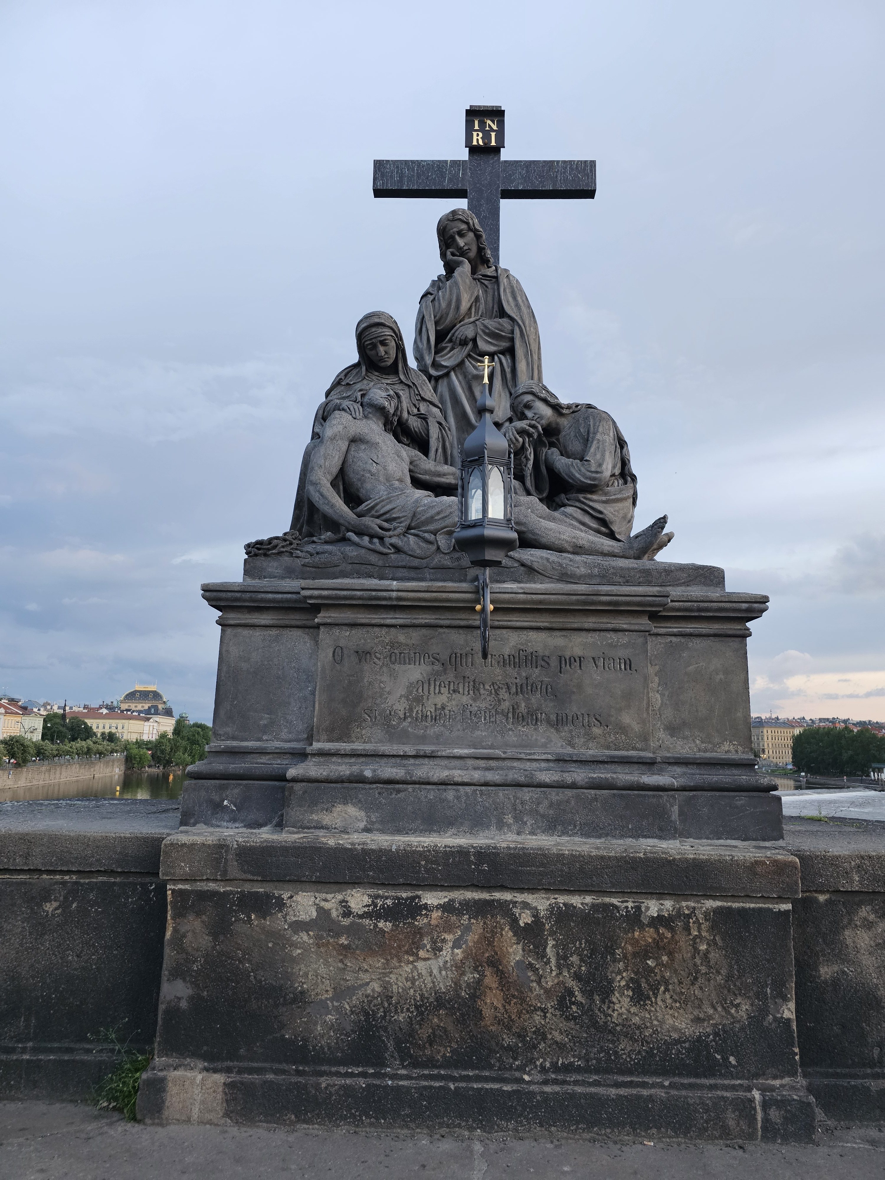 The statue of Pietà on the Charles Bridge in Prague depicts the Virgin Mary cradling the deceased Jesus Christ, with other figures present. The statue is made of dark stone and stands on a pedestal with an inscription, set against a backdrop of a cloudy sky and the city skyline.