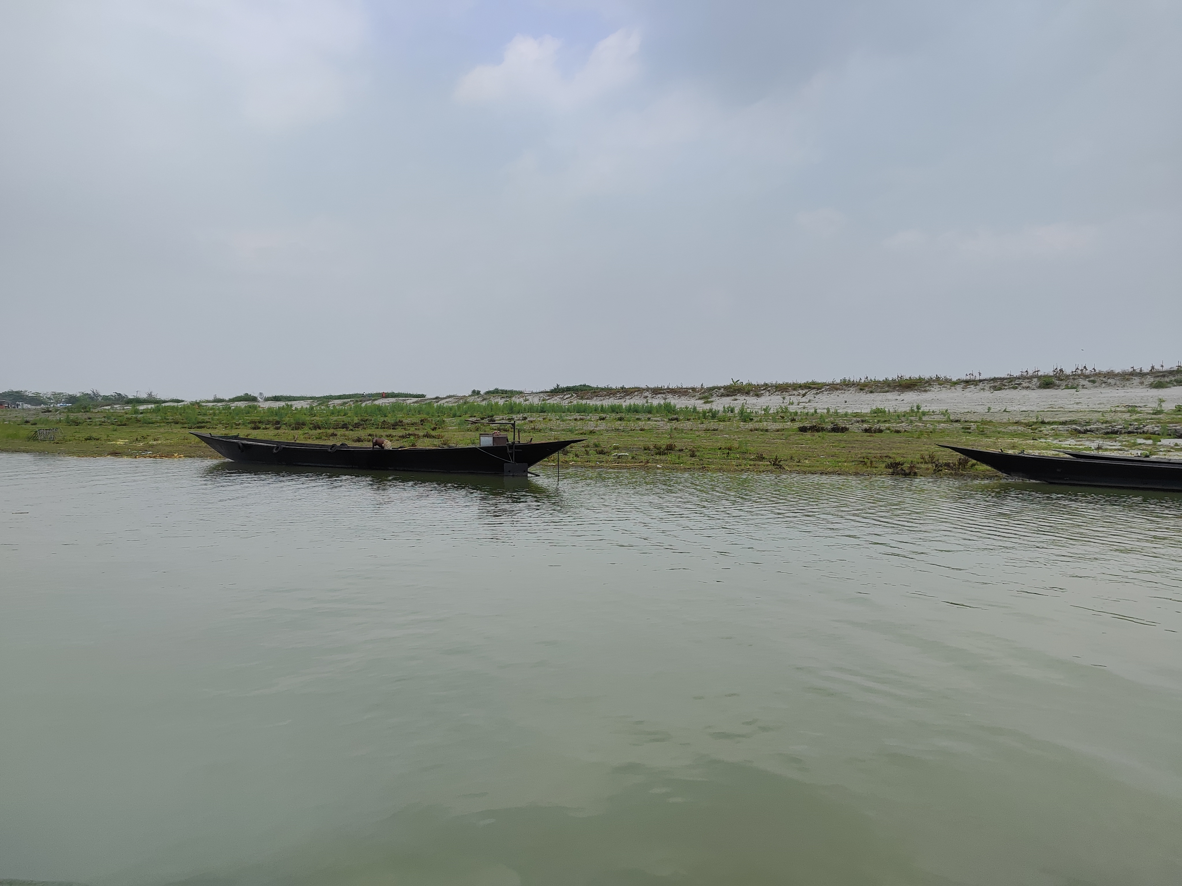 A calm river with two long, narrow boats near a grassy riverbank under a partly cloudy sky.