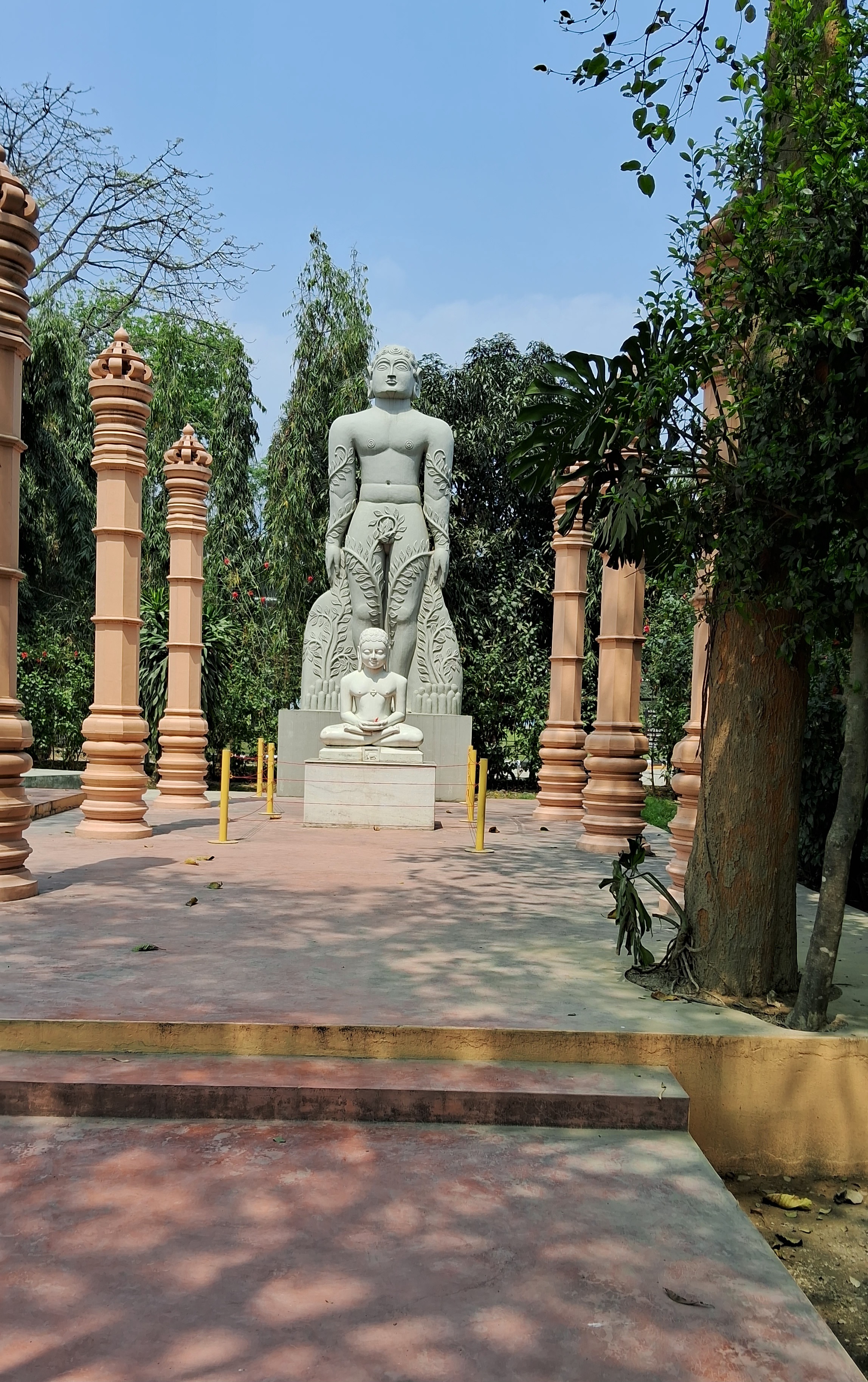 A large stone statue set amidst tall pillars and surrounded by lush greenery, with a smaller statue placed in front of it on a raised platform.