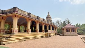 Traditional Hindu temple with yellow and red decorative architecture, open pillared hall, and a dome-shaped sanctum tower, set in a peaceful courtyard surrounded by greenery under a clear blue sky.