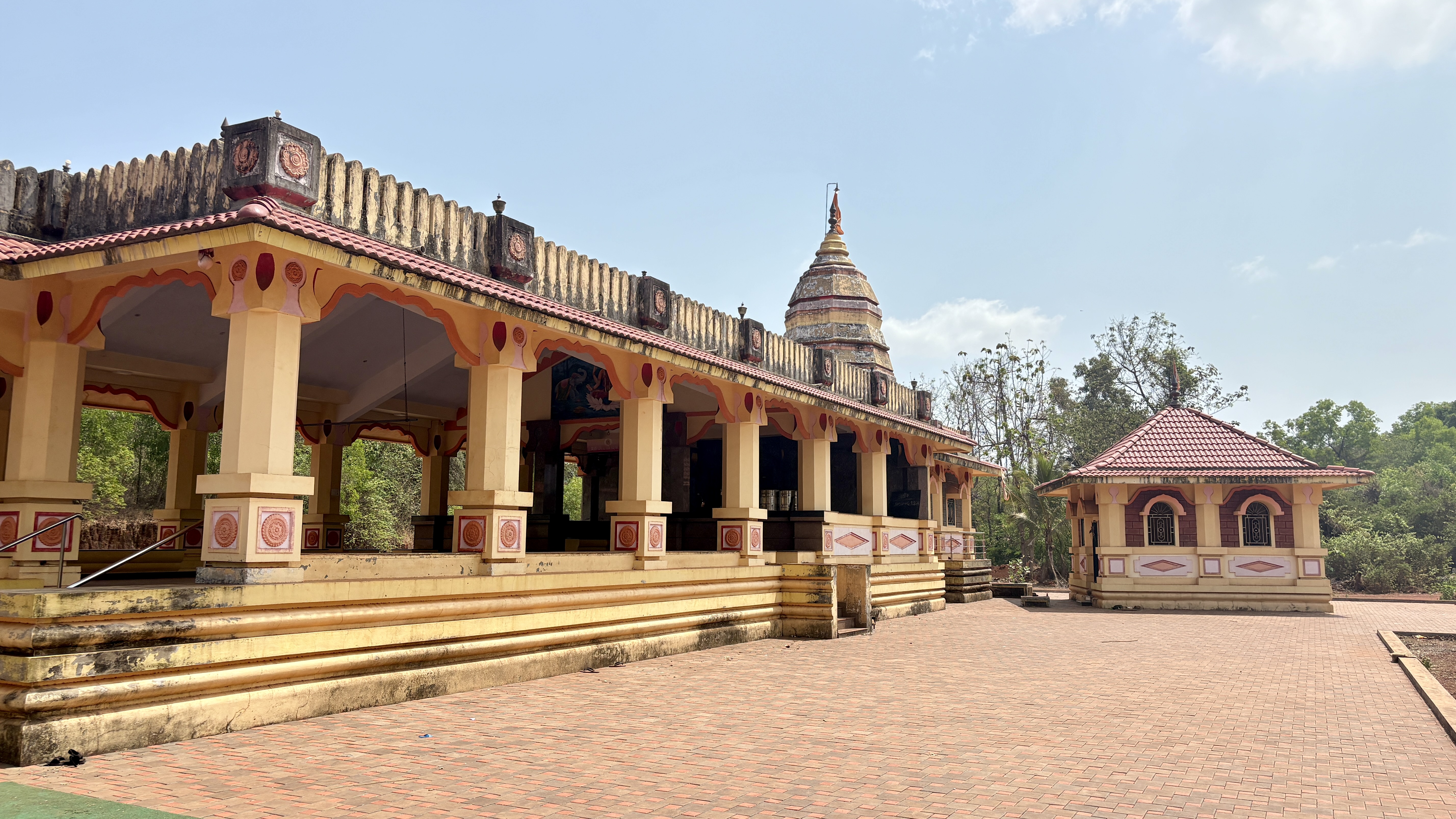 Traditional Hindu temple with yellow and red decorative architecture, open pillared hall, and a dome-shaped sanctum tower, set in a peaceful courtyard surrounded by greenery under a clear blue sky.