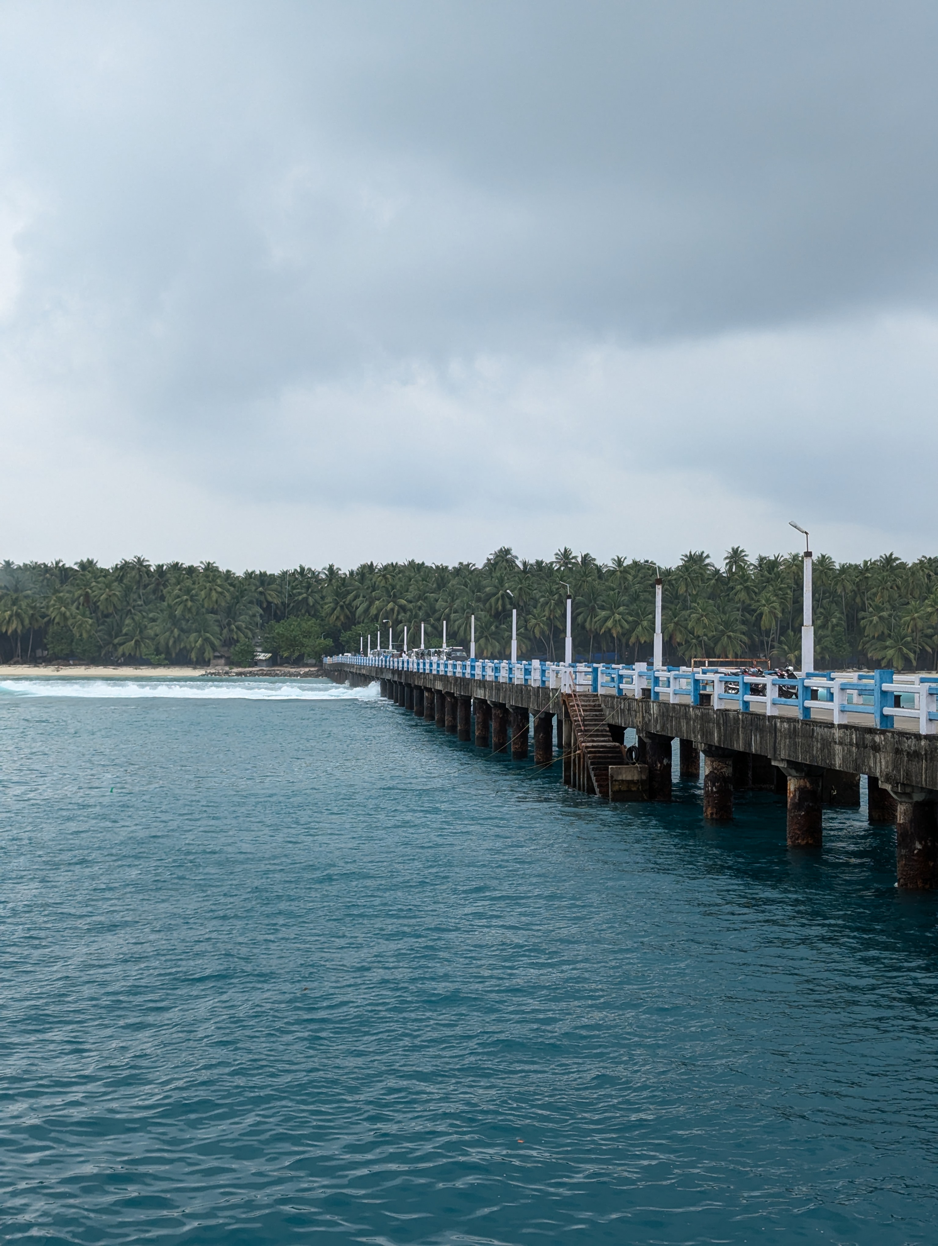 A long pier extends into calm blue waters, lined with white posts and a blue railing. In the background, lush green coconut trees rise against a backdrop of overcast skies. Waves gently break on the shore nearby, creating a tranquil coastal scene at Lakshadweep.