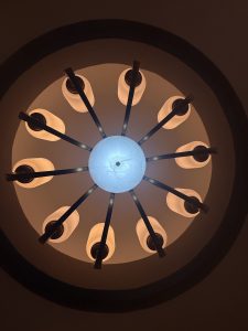 A circular chandelier with a central blue light surrounded by eight soft white lights, viewed from below against a dark background.
