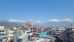 A panoramic view of a city landscape with colorful buildings in the foreground, against a backdrop of majestic snow-capped mountains under a clear blue sky. The buildings feature a variety of architectural styles and colors, while the mountains tower in the distance, showcasing their rugged peaks and glistening snow.