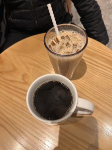 A close-up view of a wooden table featuring two beverages. One is a white ceramic cup filled with black coffee, and the other is a clear glass with iced coffee, complete with ice cubes and a white straw.
