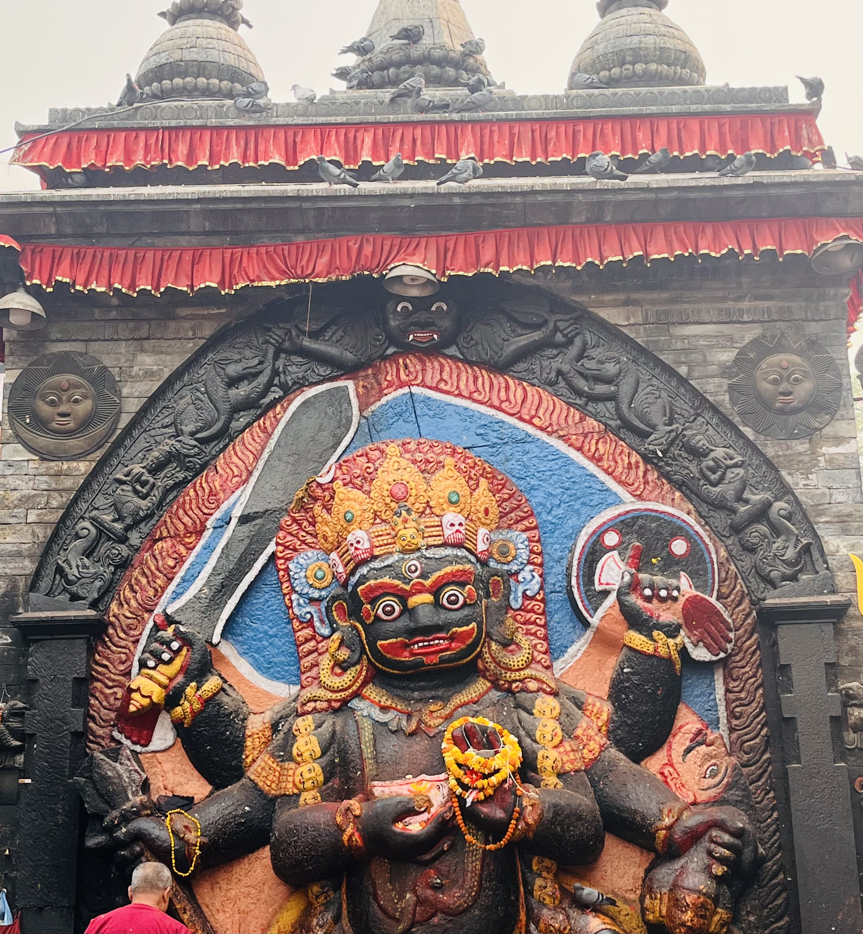 A vibrant and intricate stone sculpture of a multi-armed deity adorned with colorful ornaments, set against a decorated backdrop with red drapery