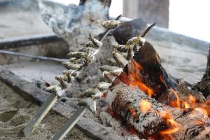 Two skewers with sardine, grilling over an open flame surrounded by ashes and sand. Smoke rises in the background, and charred logs are visible near the fire.