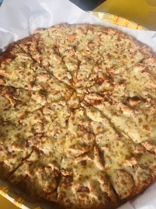 A close-up view of a large pizza topped with cheese and chicken pieces, cut into multiple slices, with a golden-brown crust. The pizza is placed on white parchment paper, with part of the box visible in the background.