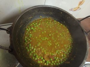 A pot of curry with green peas floating in a rich, orange-brown sauce, on a stove with a tiled kitchen backdrop.
