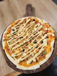 A fusion pizza, topped with chicken, green peppers, onions, and black olives, drizzled with a creamy sauce. It is placed on a wooden board. Spotted it at a restaurant in Kozhikode, Kerala. #WPNP2025
