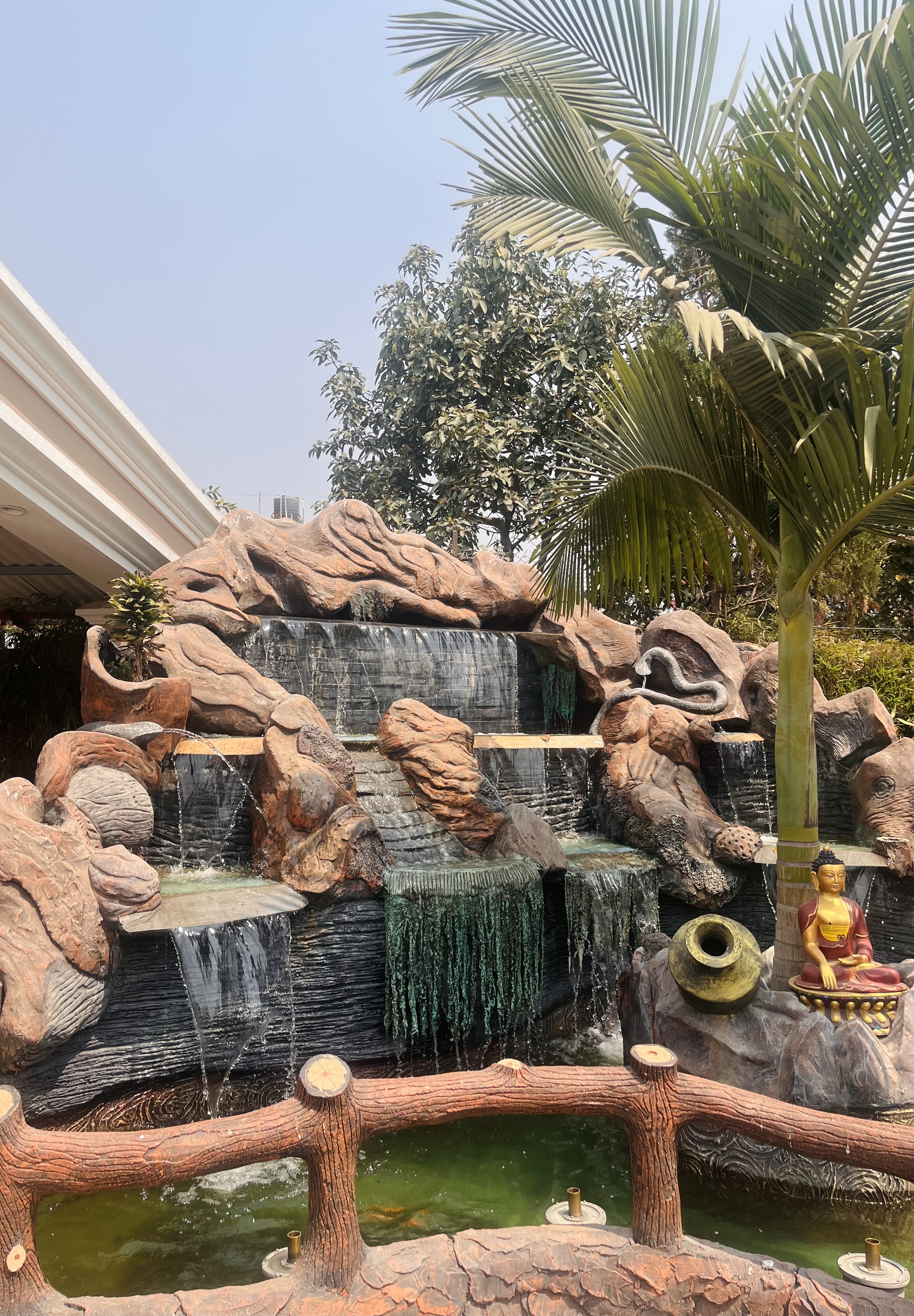 A decorative outdoor waterfall with multiple tiers of cascading water, surrounded by artificial rocks. There is a statue of a seated figure next to an urn, and a palm tree is visible on the right. A fence resembling tree branches is in the foreground, with green foliage and a clear sky in the background.