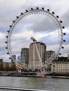 A large Ferris wheel by a river with modern and historic buildings in the background, under a cloudy sky.