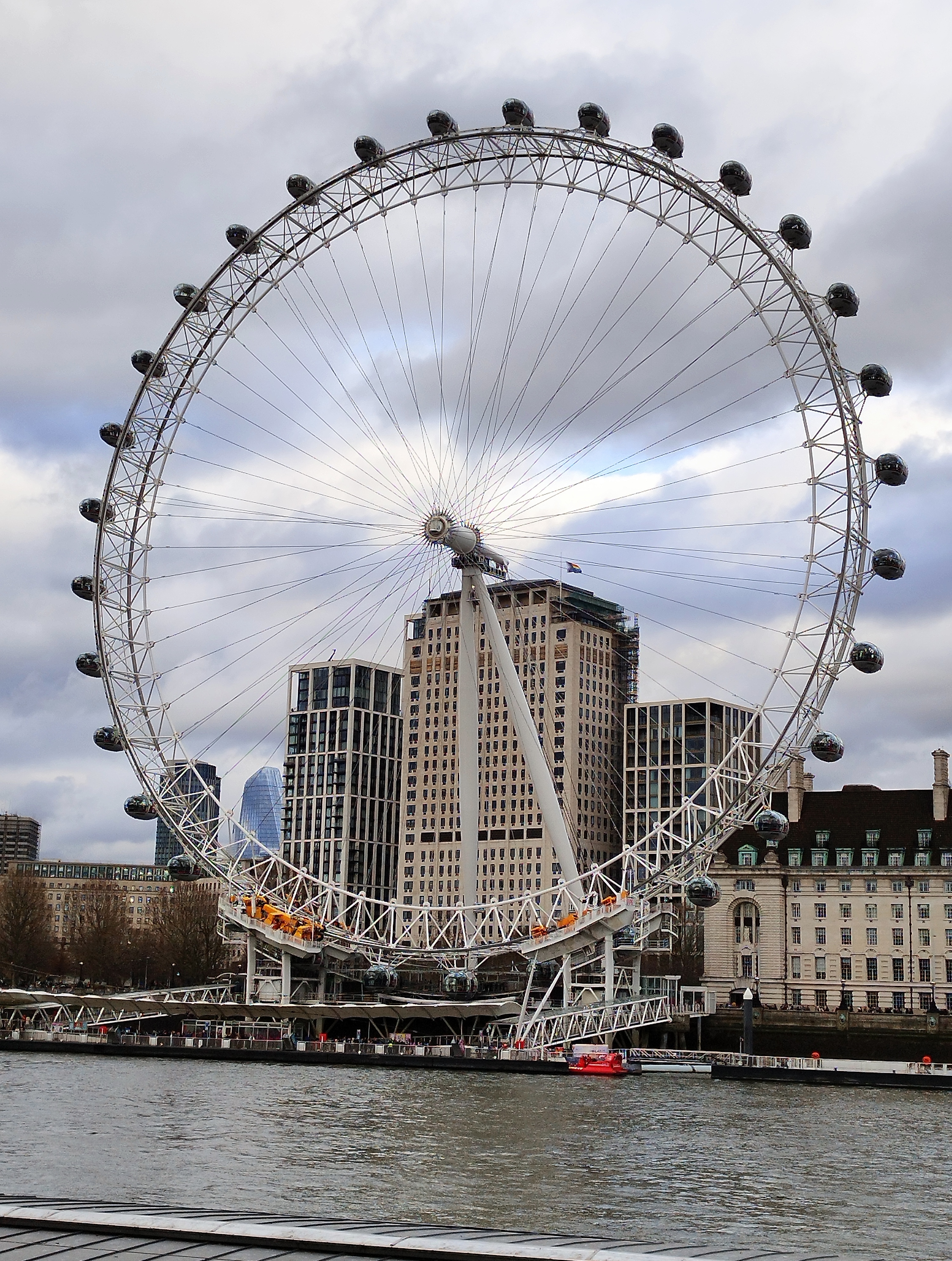 A large Ferris wheel by a river with modern and historic buildings in the background, under a cloudy sky.