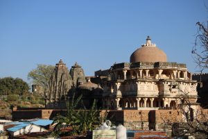 Ancient stone temple complex with intricate carvings and a large dome, set against a clear blue sky. Vegetation and smaller structures are visible in the foreground.