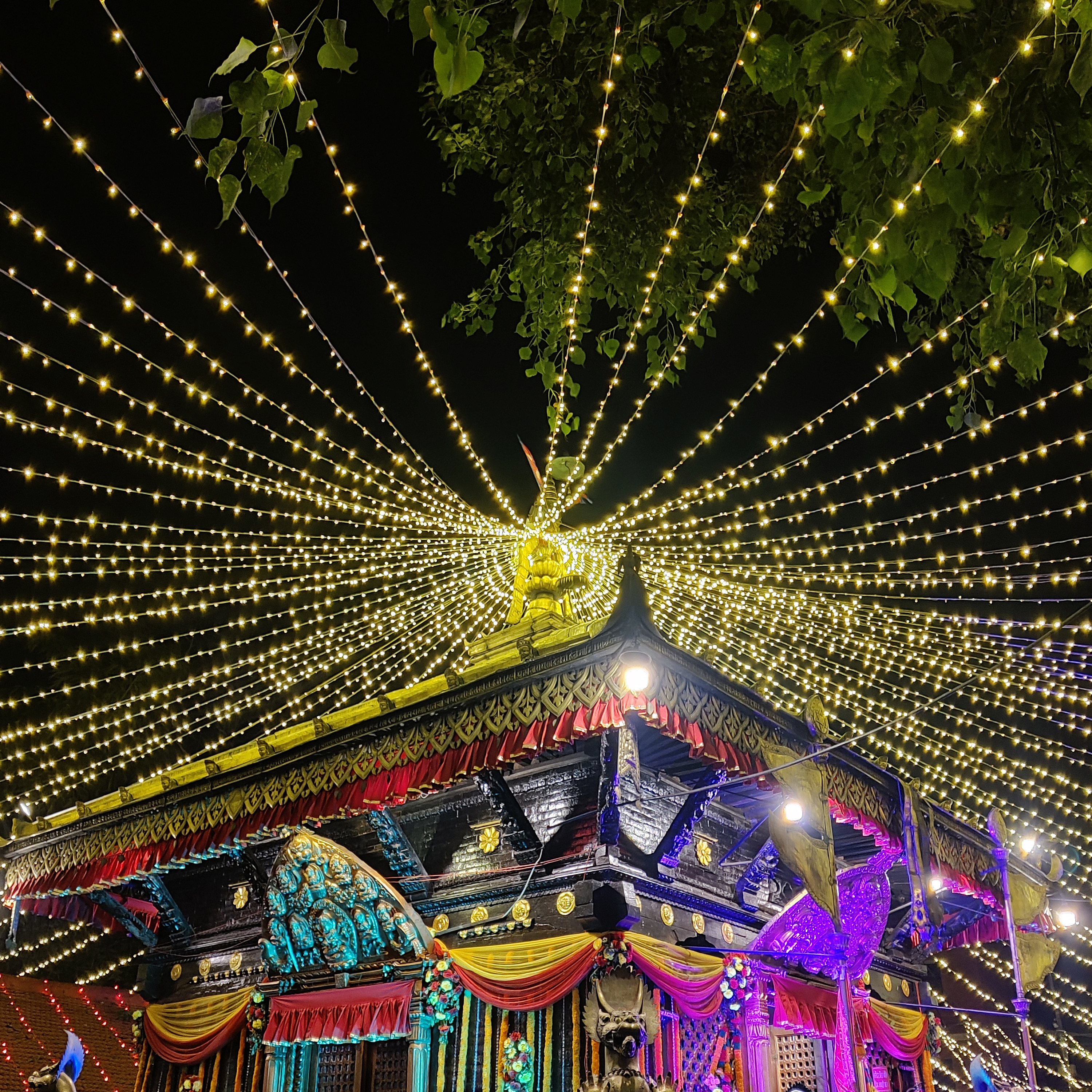 An intricately decorated temple illuminated with colorful lights. Strings of small, bright lights radiate outward from the temple's spire against the night sky.