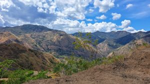 A scenic view of rugged mountains under a partially cloudy sky. The landscape features a mix of green foliage and exposed rock, with a tree in the foreground. The clouds are scattered, allowing patches of blue sky to be visible.