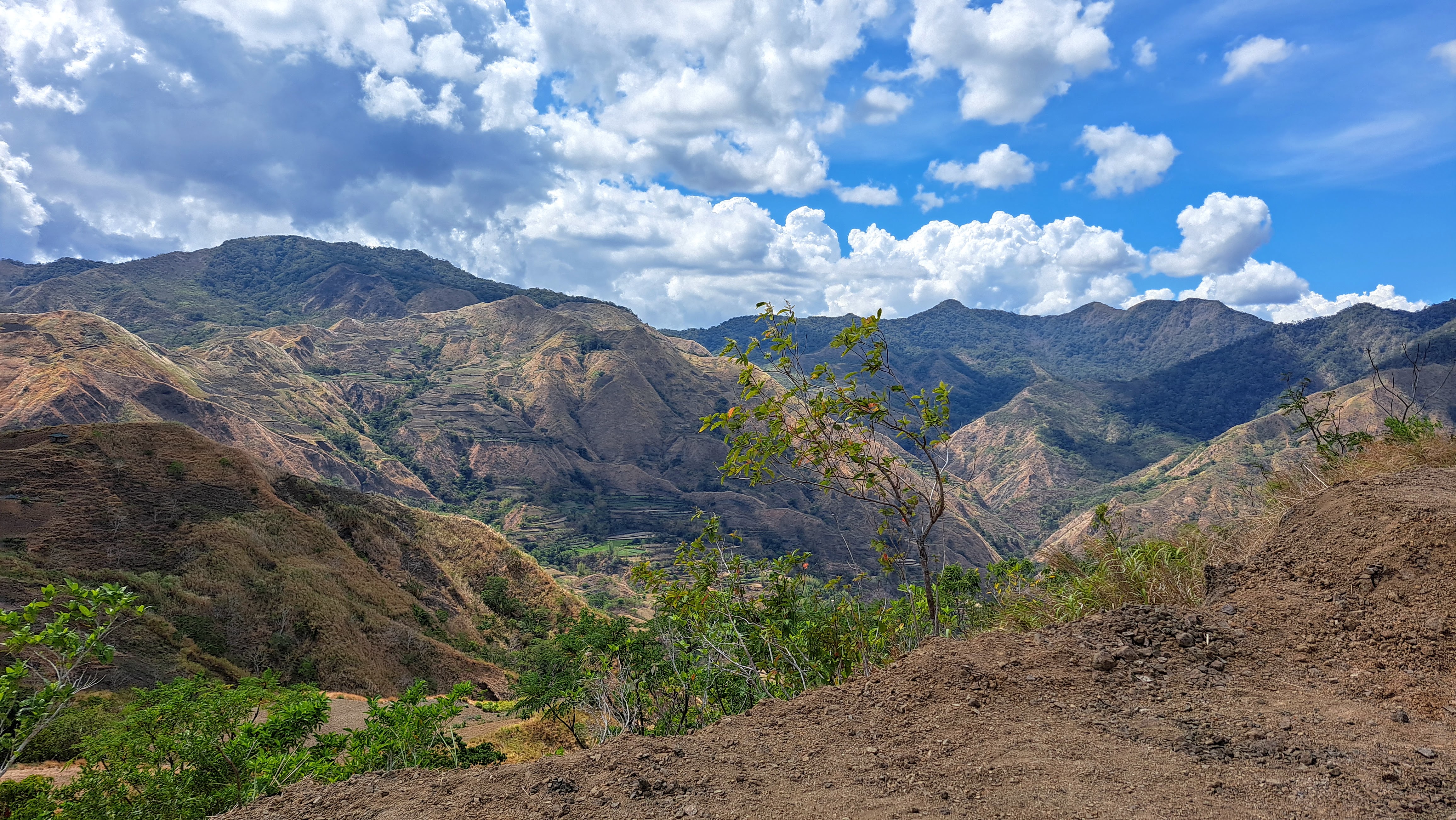 A scenic view of rugged mountains under a partially cloudy sky. The landscape features a mix of green foliage and exposed rock, with a tree in the foreground. The clouds are scattered, allowing patches of blue sky to be visible.