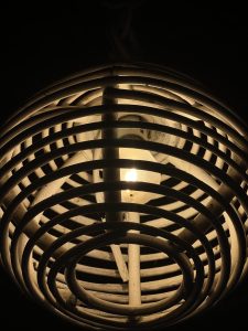 A close-up of a pendant light fixture with a cage-like design made of thin, round metal strips. The light bulb inside emits a warm glow, illuminating the surrounding area while the background remains dark. 