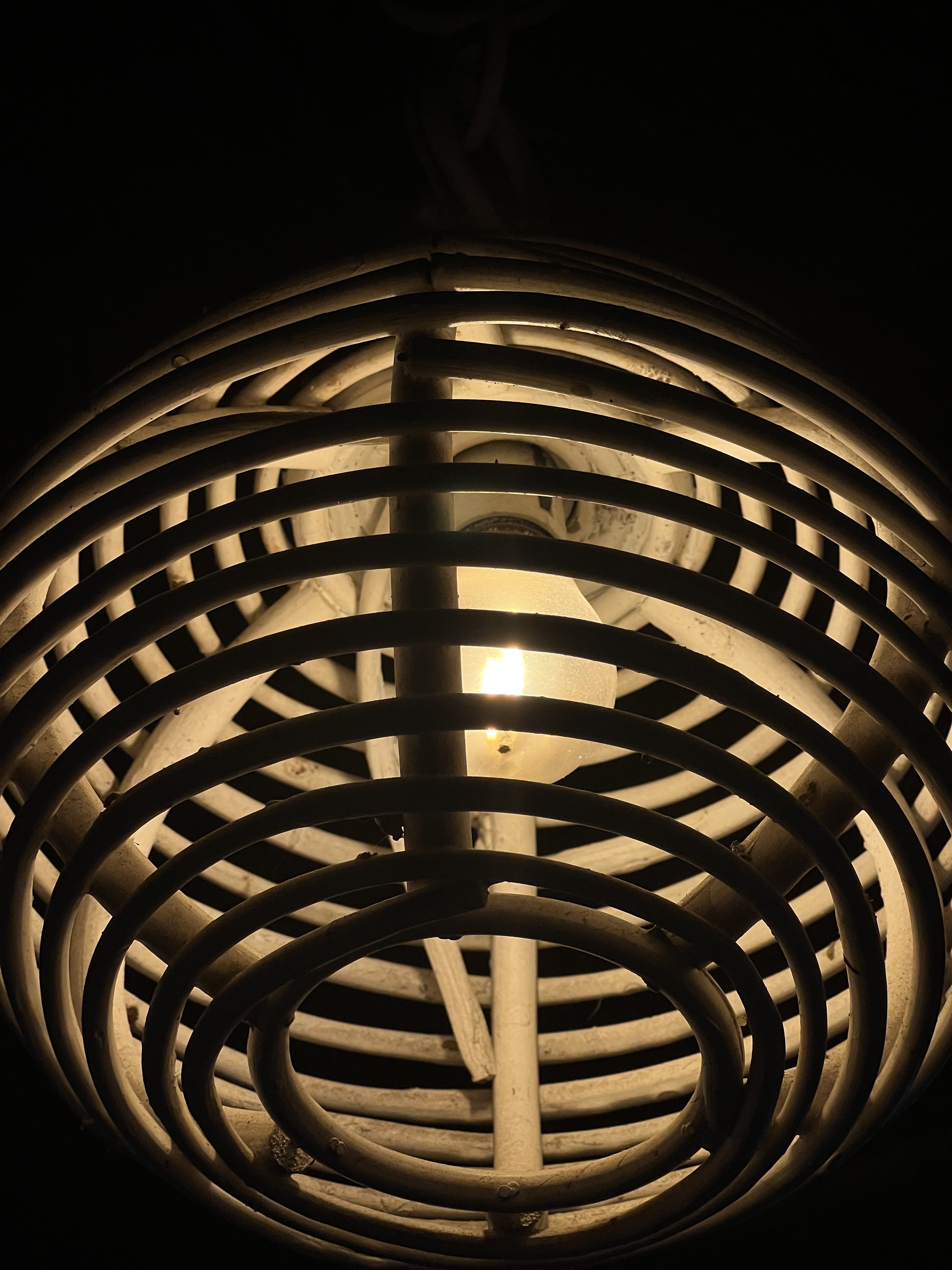 A close-up of a pendant light fixture with a cage-like design made of thin, round metal strips. The light bulb inside emits a warm glow, illuminating the surrounding area while the background remains dark.