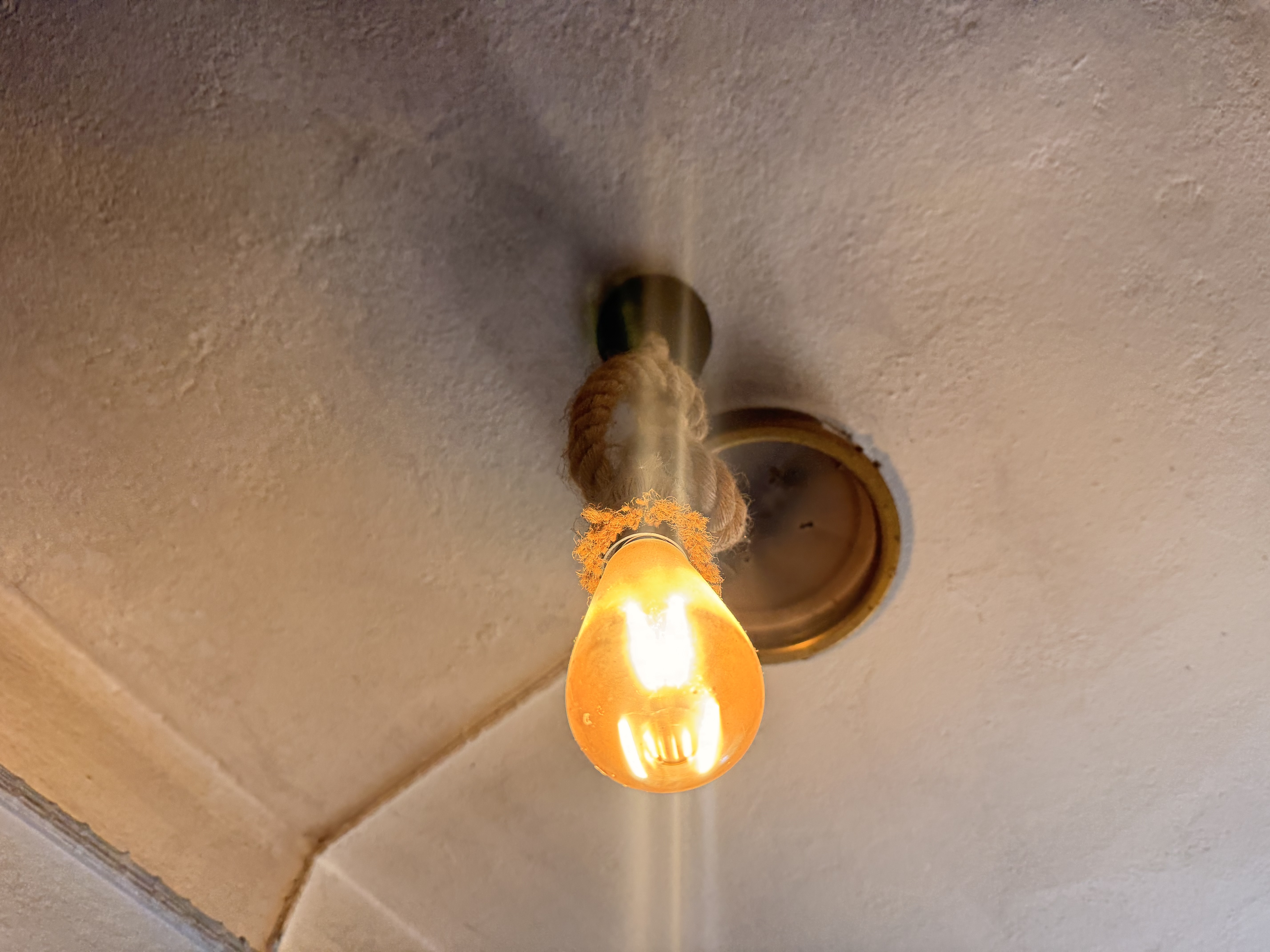 An illuminated light bulb hanging from a ceiling fixture, attached with a rope, against a textured ceiling.