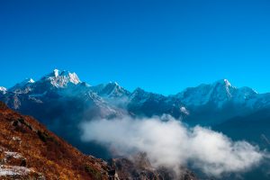 A breathtaking view of a mountain range with snow-capped peaks under a clear blue sky. In the foreground, a slope covered in brown grass and a layer of mist or clouds partially covers the landscape.