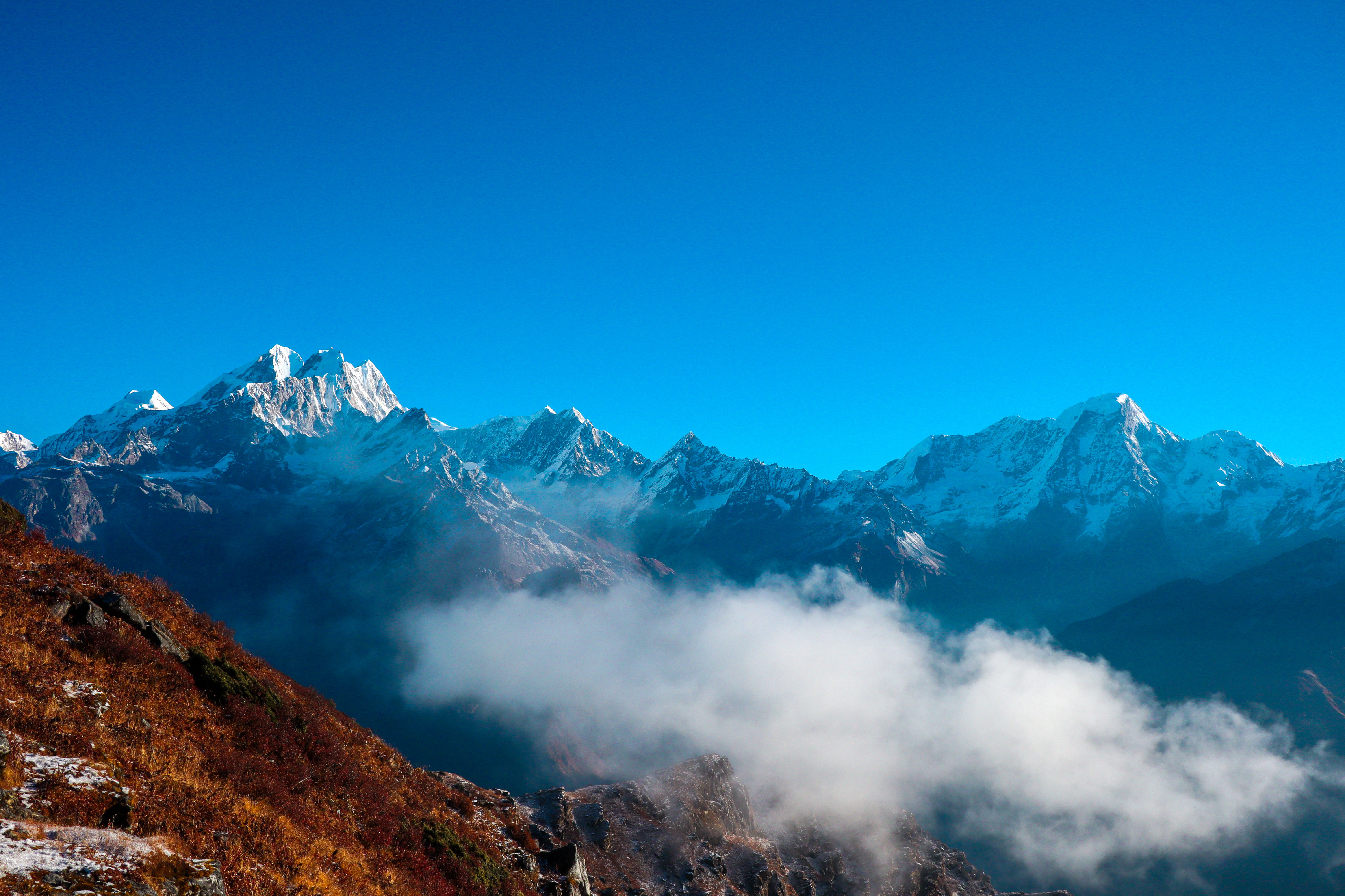 A breathtaking view of a mountain range with snow-capped peaks under a clear blue sky. In the foreground, a slope covered in brown grass and a layer of mist or clouds partially covers the landscape.