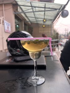 A close-up of a cocktail glass filled with a light-colored drink, garnished with mint leaves, and a pink straw resting on the rim. 