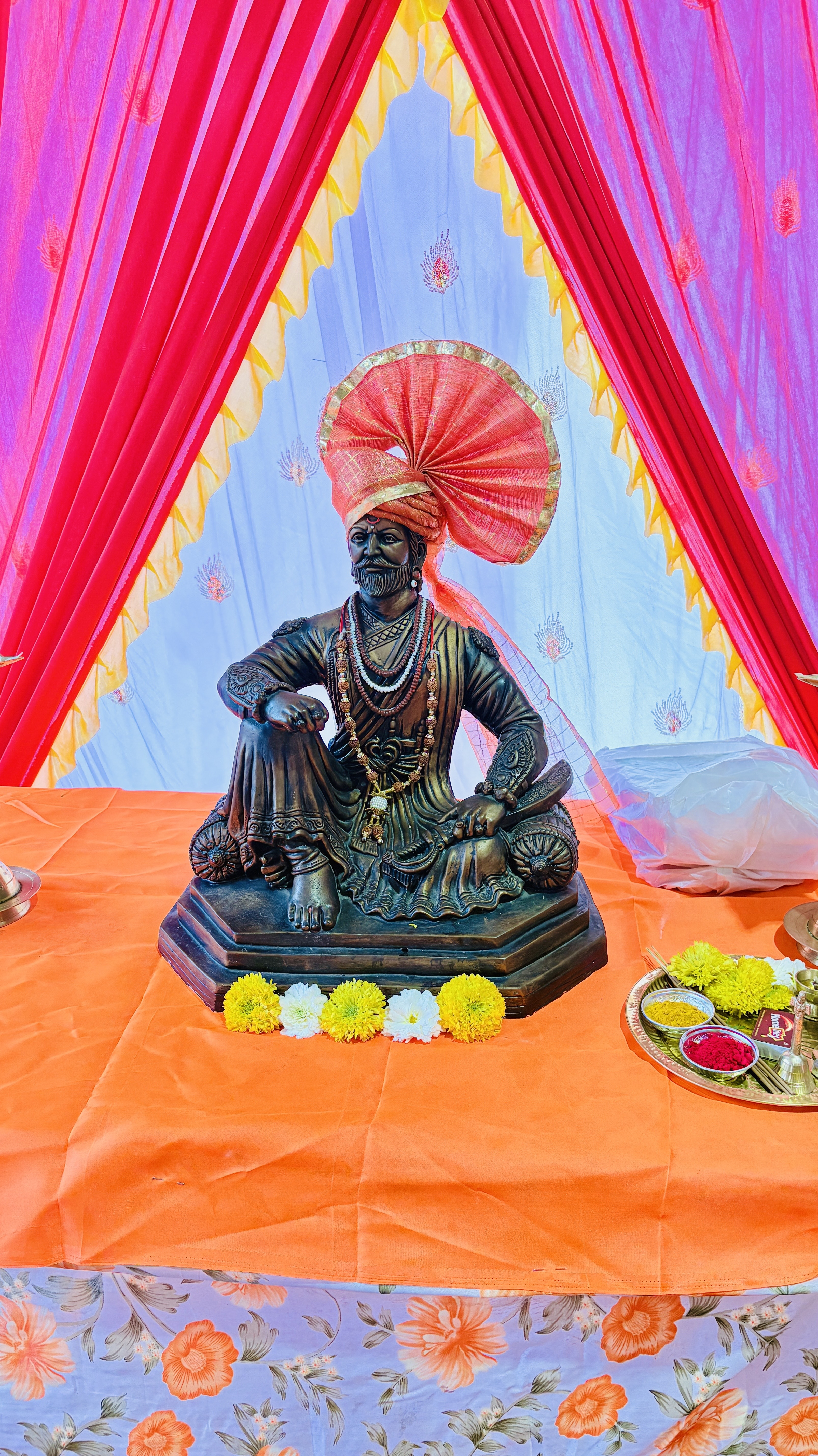 A statue of Chhatrapati Shivaji Maharaj, a revered historical figure, is placed on an orange cloth-covered table. The statue depicts him seated, wearing traditional attire with a turban adorned with a decorative fan-like ornament.