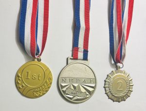 Three medals with red, white, and blue ribbons are displayed: a gold medal labeled "1st," a silver medal with "NEPAL" and a map of the country, and a bronze-toned medal labeled "2."