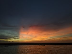 Dramatic sunset over water, dark sky above transitioning to orange and yellow hues, with a distant boat silhouette
